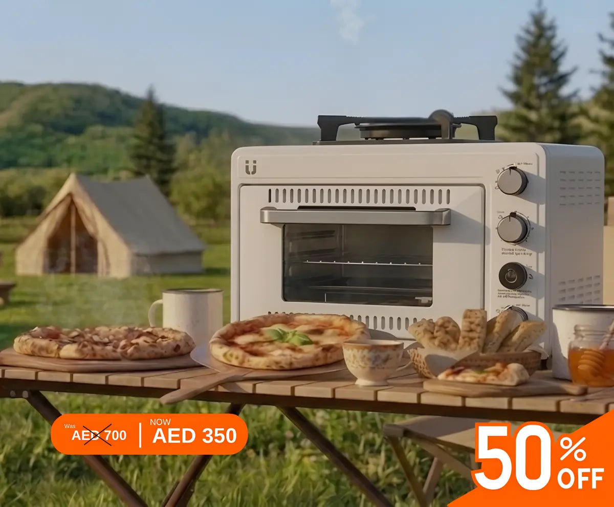 Picnic table with portable oven and stove, pizza, and drinks in a scenic outdoor setting with mountains and trees.